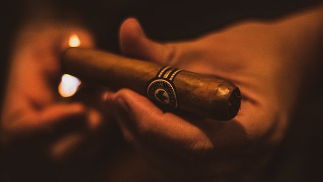 Hands holding a lit premium cigar with a decorative band in warm amber lighting at a cigar lounge